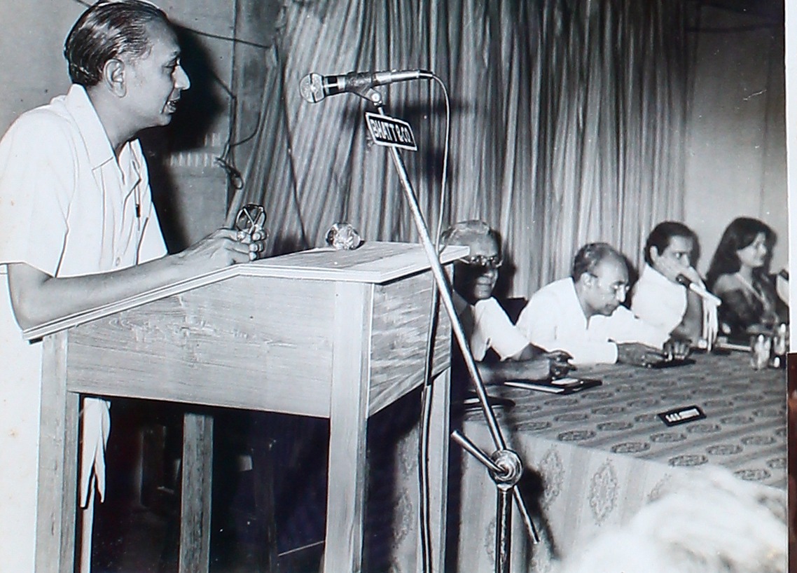 A man addressing a gathering, a panel seated alongside him.